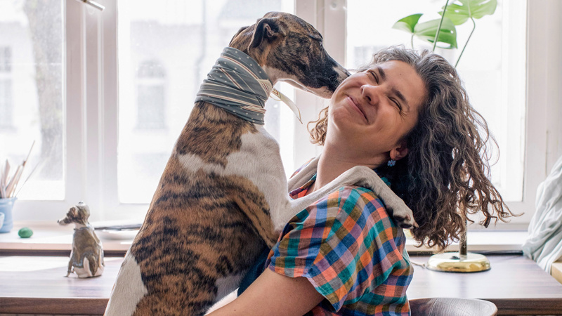 Kvinna lyfter upp stor hund i ljust rum med soffa – lekfull bild.