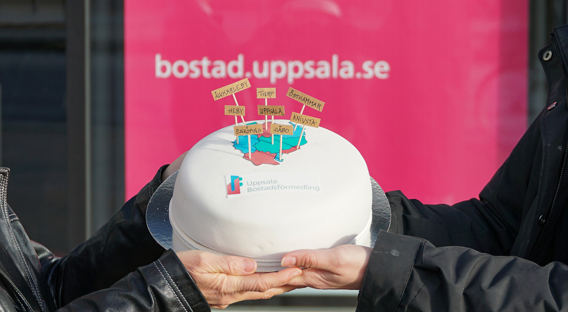 Person håller likadan tårta med texten "bostad.uppsala.se" i bakgrunden.